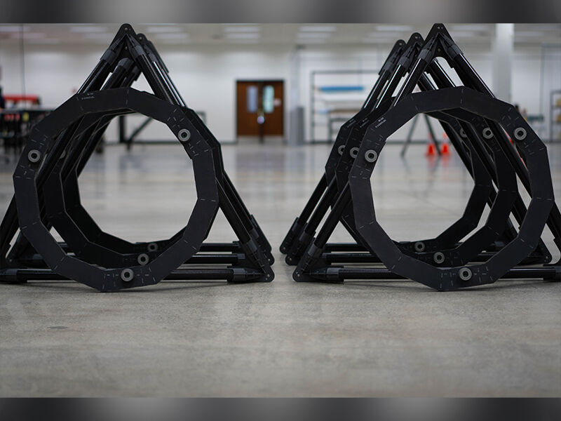 Image of TMT metrology fixtures lined up on the floor of a manufacturing area.  There are two rows of three and the viewer is at eye level to the precision fixtures. The hardware appears as black circular shapes with a hollow center that are attached at three major points to black triangular shapes.